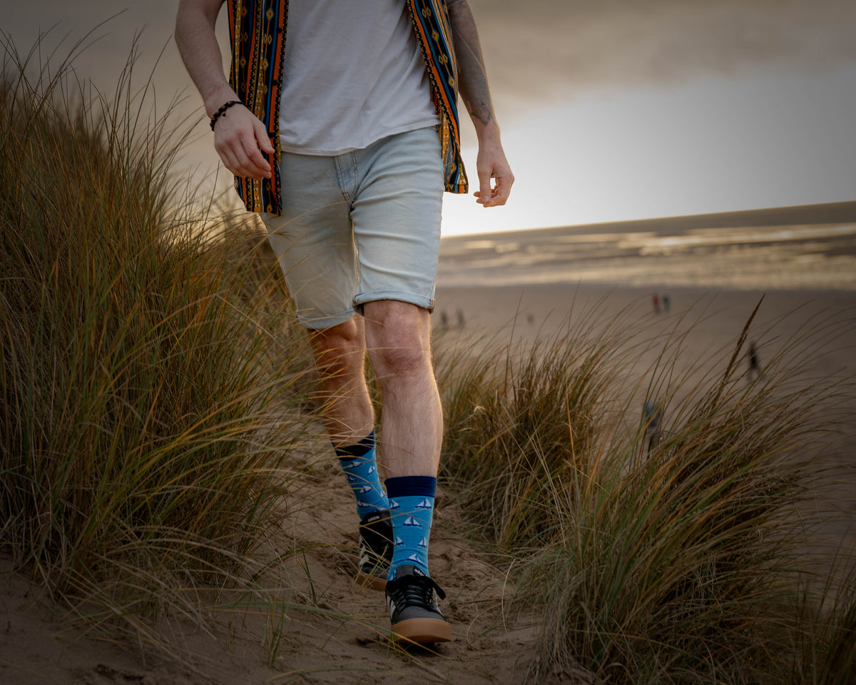 Man walking a sandy dune path through beach grass, wearing blue sailing boat bamboo socks with navy cuffs and black trainers